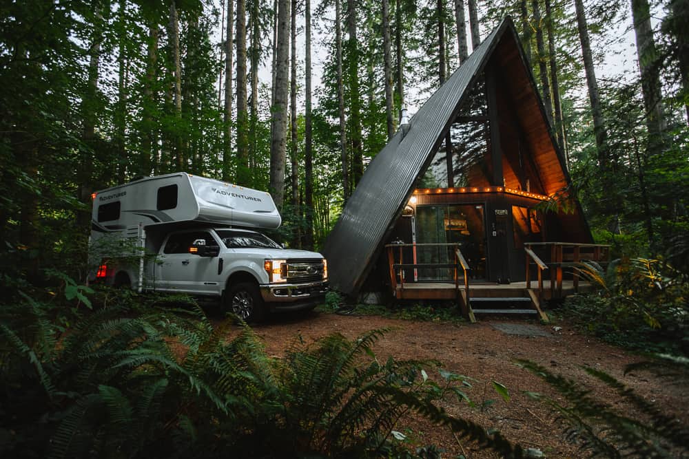 A truck camper (the adventurer) on the back of a pickup truck next to an A Frame house in the forest