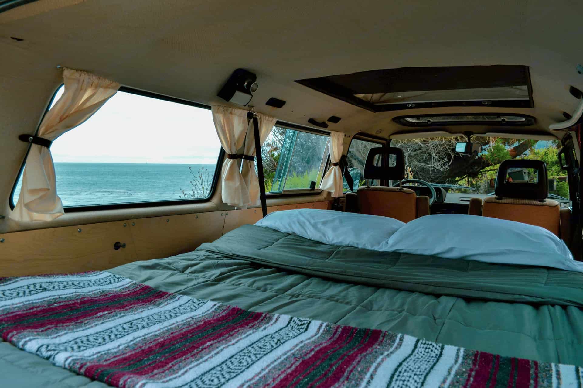 A made bed in the back of a VW camper van with the ocean through the open door