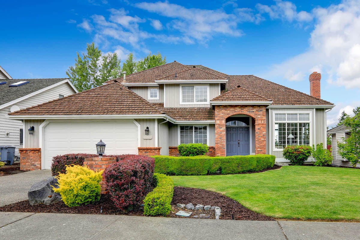 A mid-size house with a brick entryway and a garage. There is green grass in the front and colorful bushes.