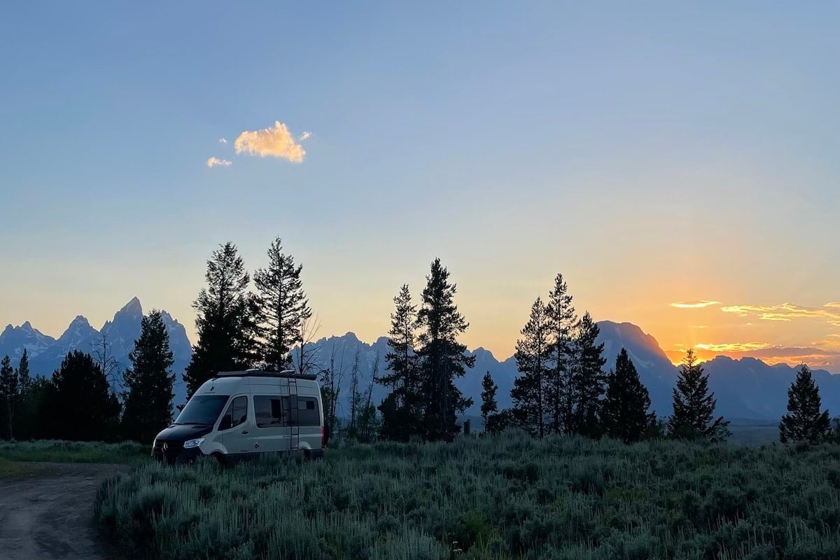 A photo of a van with mountains and trees in the background at sunset.