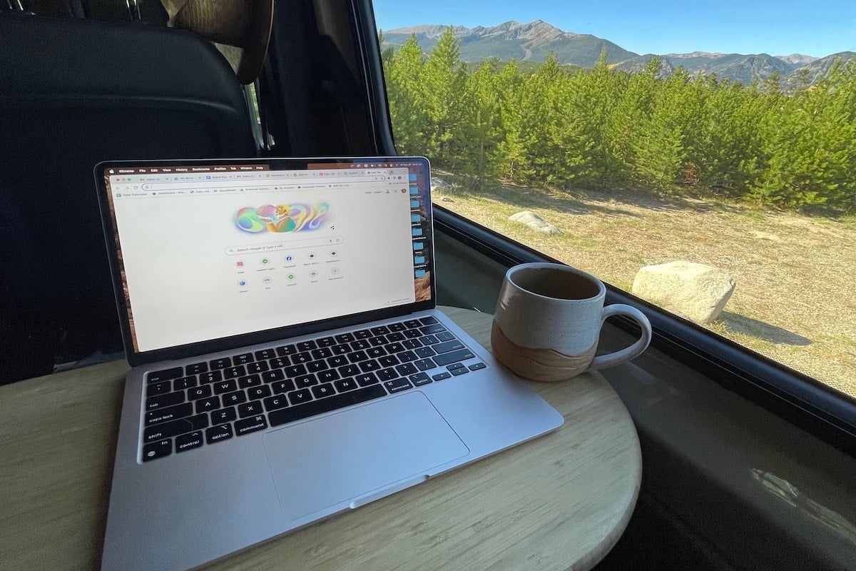 Photo of a laptop with a coffee mug next to it and the view of mountains with a blue sky out of the window of a cargo van.