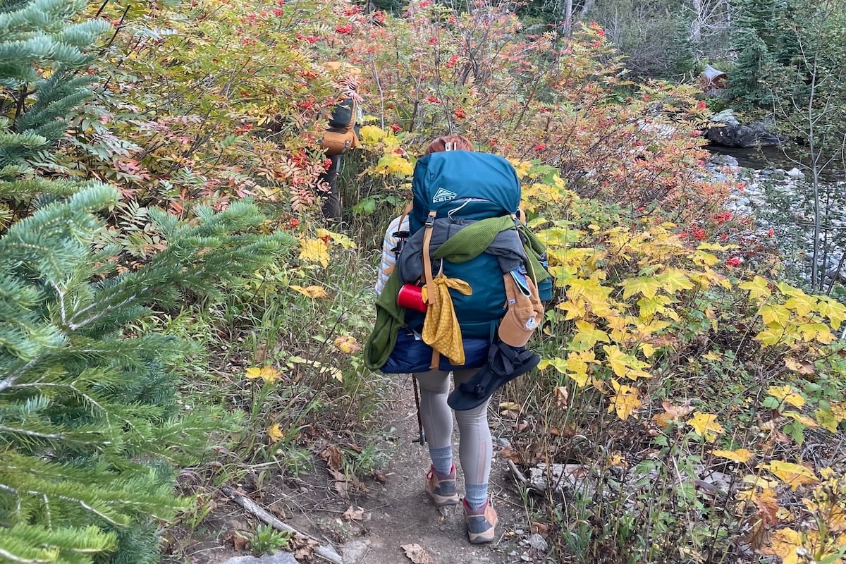 Photo of a woman with a teal backpacking pack with items attached to it hiking on a dirt trail with yellow leaves.
