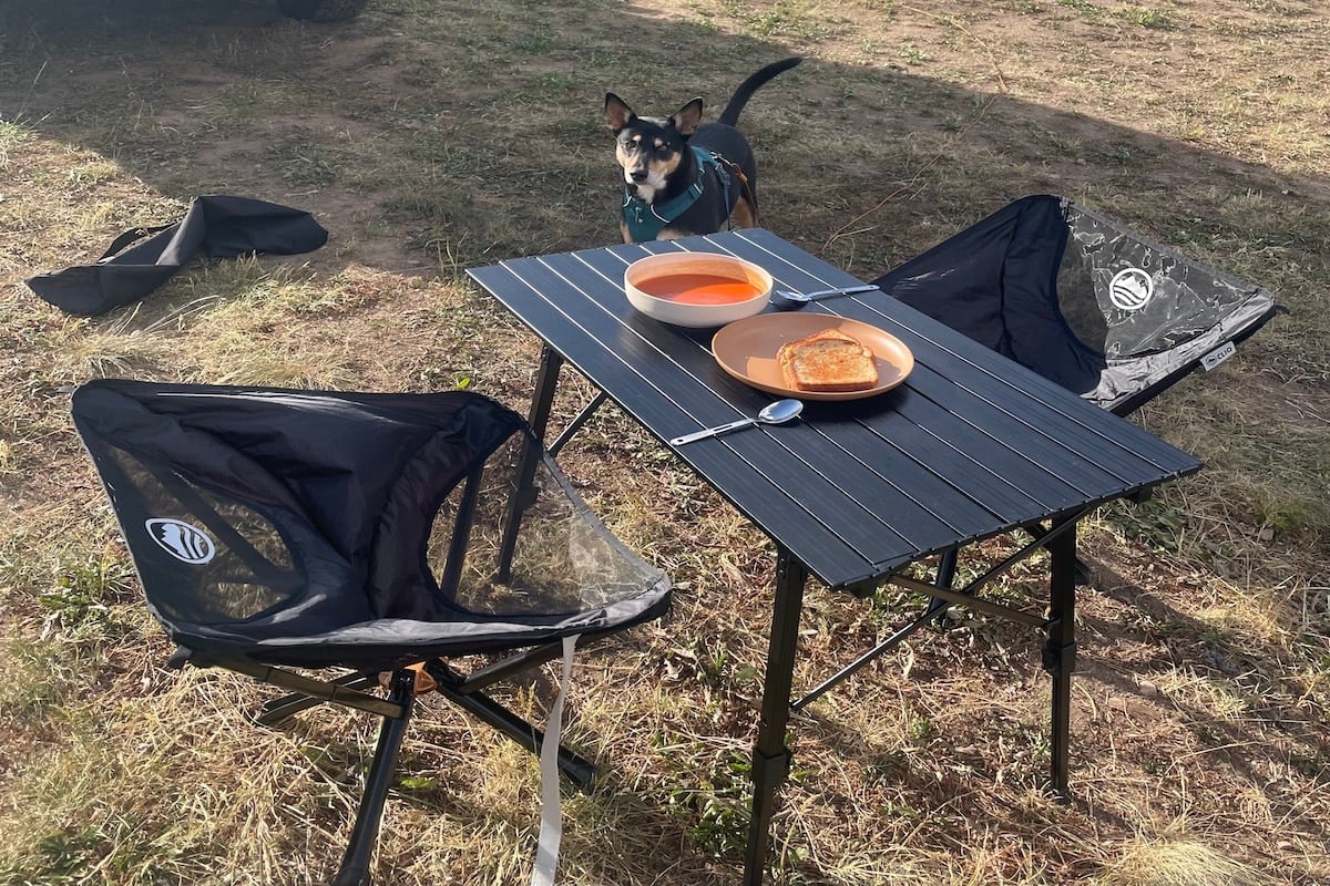 Two black camp chairs with a black camping table that has a bowl of tomato soup and a sandwich on it. There is a black dog and grass in the background.