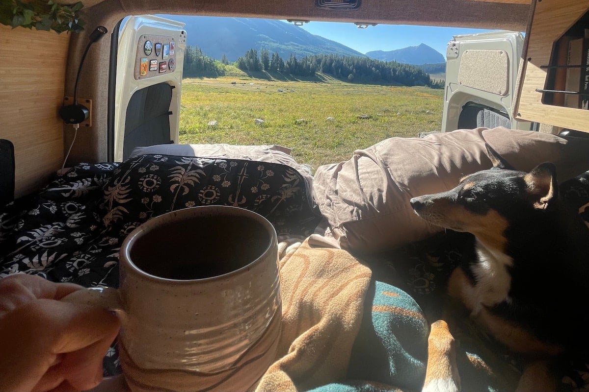 A hand holding a coffee mug with coffee in it. There is a dog, blanket, and pillows. The back two doors of a van are open to green grass and mountains outside.