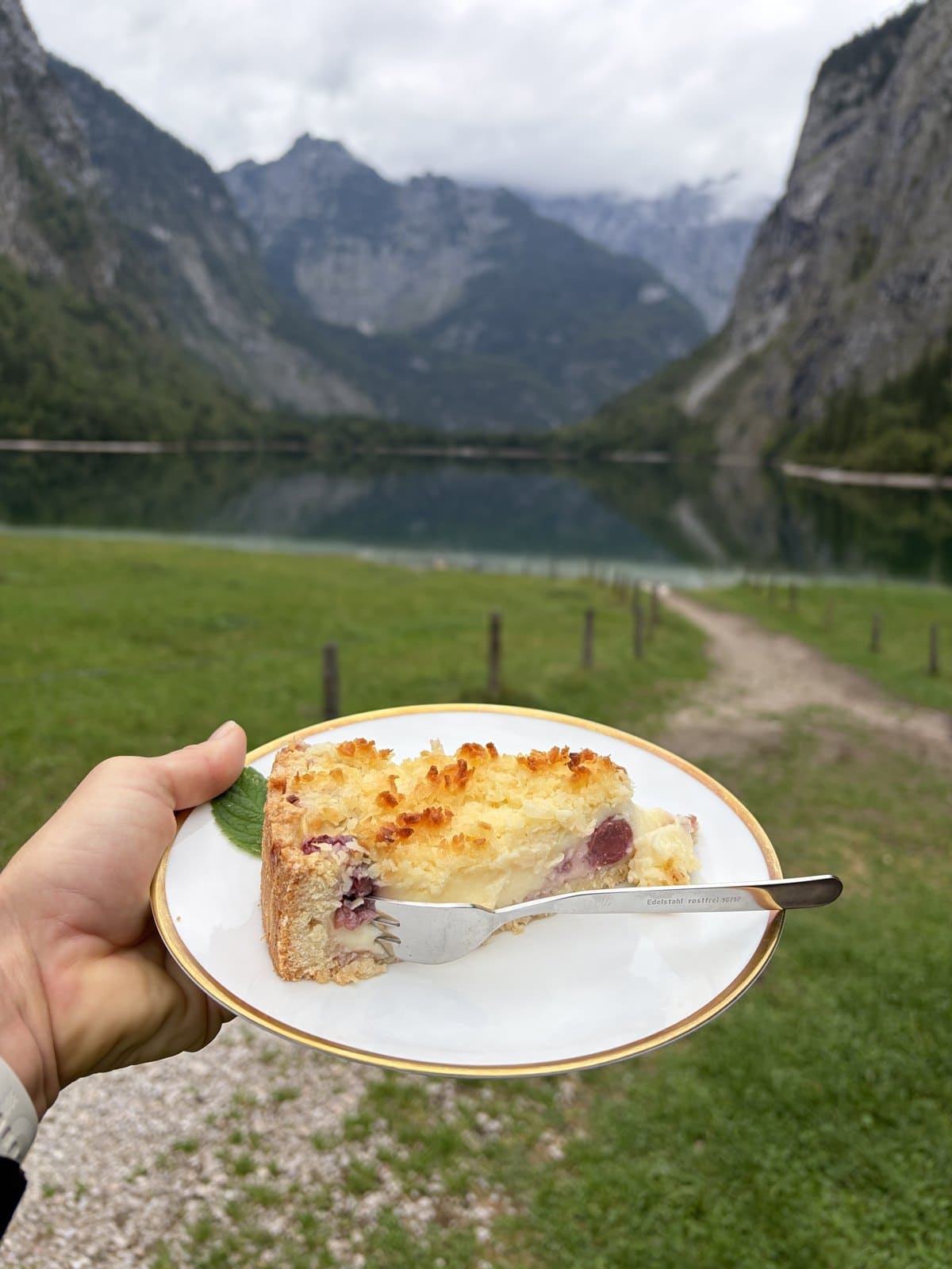 A slice of cherry cheesecake on a plate held up in front of the Obersee and its mountain hut