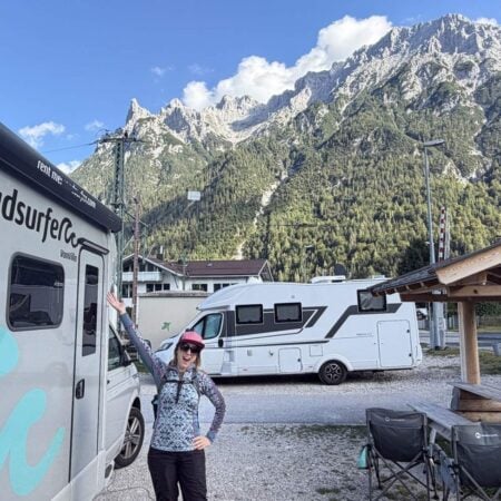 Kristin with her arms out beside a Roadsurfer Van Villa campervan at a camping area in Mittenwald, the Karwendel mountains behind
