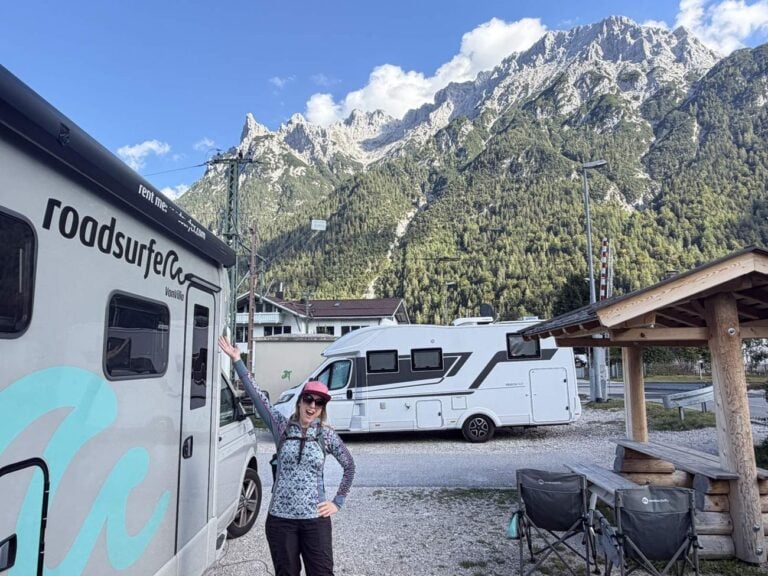 Kristin with her arms out beside a Roadsurfer Van Villa campervan at a camping area in Mittenwald, the Karwendel mountains behind