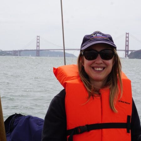 Kristin in an orange life jacket and navy cap sailing on San Francisco Bay with the Golden Gate Bridge behind her