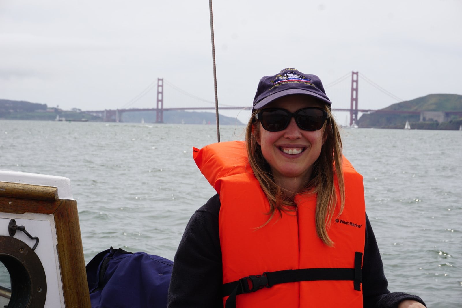 Kristin in an orange life jacket and navy cap sailing on San Francisco Bay with the Golden Gate Bridge behind her