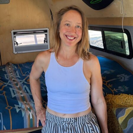 Kristin smiling inside her Sprinter campervan, standing beside the bed with the plywood walls and windows behind her