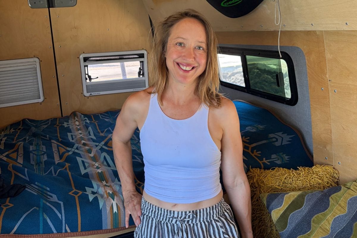 Kristin smiling inside her Sprinter campervan, standing beside the bed with the plywood walls and windows behind her