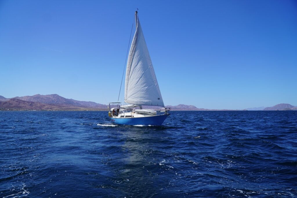 A sailboat under full sail on blue water in the Sea of Cortez with desert mountains behind