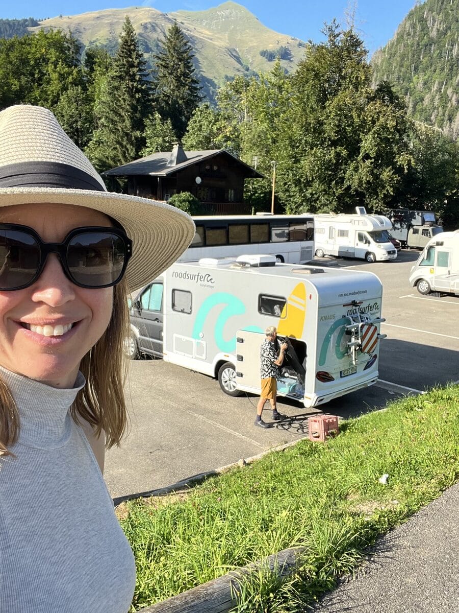 Kristin smiling in a sun hat with Tom and their Roadsurfer camper in the French Alps